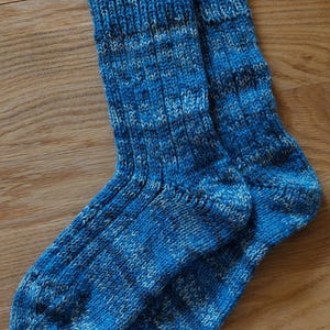 Puede incluir: Un par de calcetines tejidos a mano en tonos azules y blancos, sobre una superficie de madera. Los calcetines presentan un pu&ntilde;o acanalado y un patr&oacute;n de tejido texturizado, para mayor comodidad.