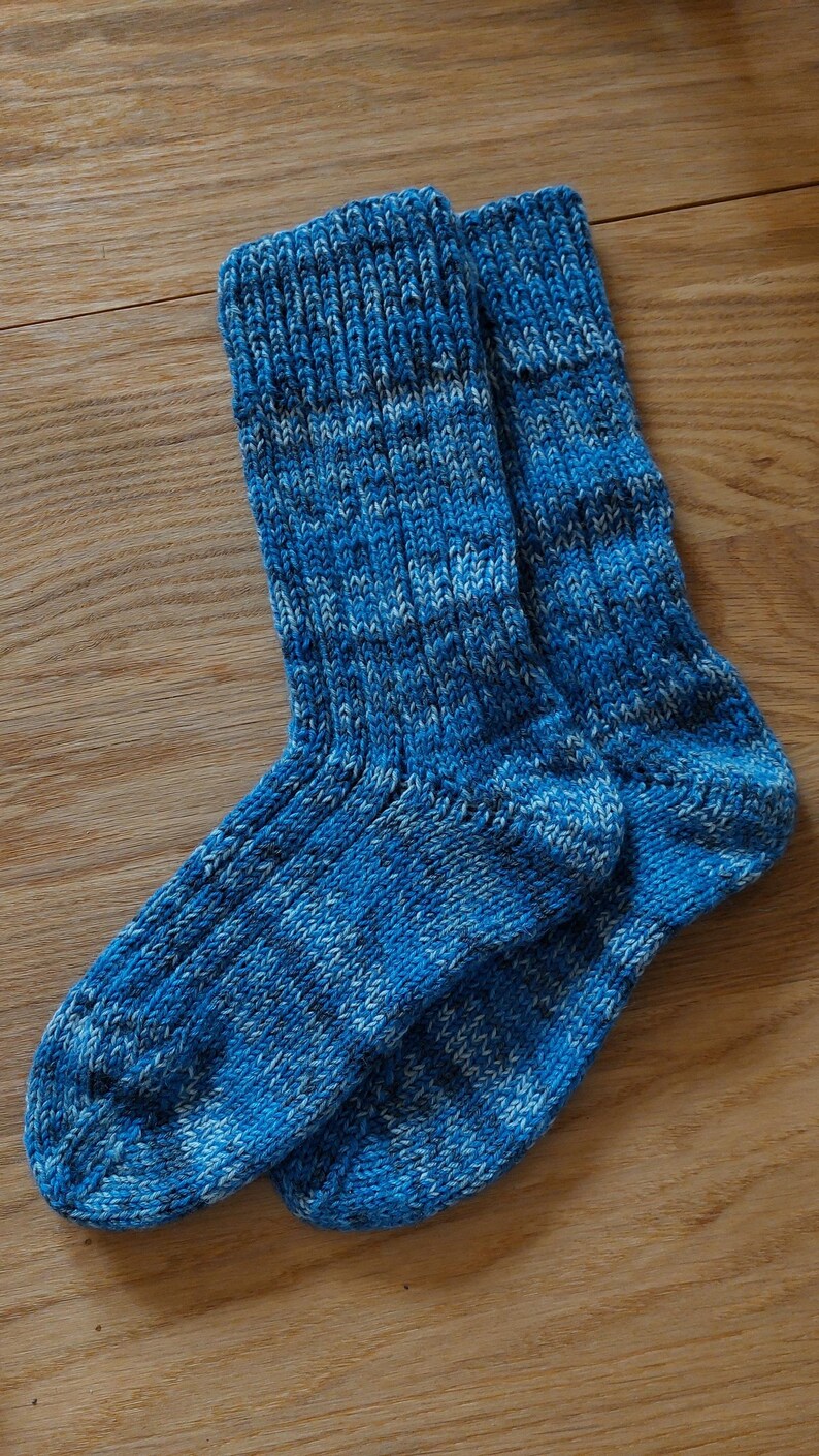Puede incluir: Un par de calcetines tejidos a mano en tonos azules y blancos, sobre una superficie de madera. Los calcetines presentan un pu&ntilde;o acanalado y un patr&oacute;n de tejido texturizado, para mayor comodidad.