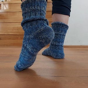 Puede incluir: Primer plano de calcetines azules de punto con pu&ntilde;o doblado. Los calcetines tienen un aspecto texturizado con una mezcla de hilos azules y blancos. Los calcetines se llevan sobre un suelo de madera, con una escalera al fondo.