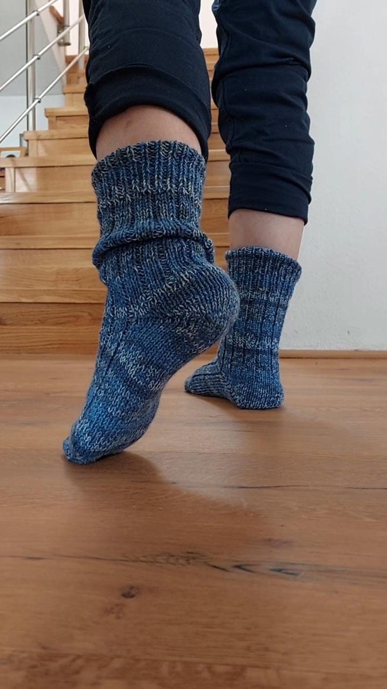 Puede incluir: Primer plano de calcetines azules de punto con pu&ntilde;o doblado. Los calcetines tienen un aspecto texturizado con una mezcla de hilos azules y blancos. Los calcetines se llevan sobre un suelo de madera, con una escalera al fondo.