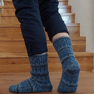 Puede incluir: Un par de calcetines de punto azules con un patr&oacute;n texturizado. Los calcetines se usan con pantalones azul oscuro, y el fondo presenta escaleras y pisos de madera.