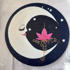 May include: A white crescent moon with a closed eye and eyelashes painted on it, with a pink lotus flower and gold swirls on a dark blue background with white stars.