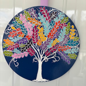 May include: A round wooden wall hanging with a white tree painted on a dark blue background. The tree has colorful leaves in shades of pink, purple, blue, green, yellow, orange, and red.