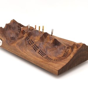 May include: A wooden game board shaped like a mountain range with a textured surface and holes. The board has five metal pegs inserted into the top of the mountain.