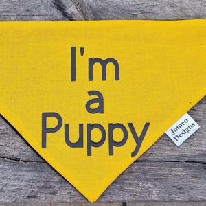 May include: Yellow dog bandana with black text that reads "I'm a Puppy" and a small white tag that reads "Jomeo Designs".