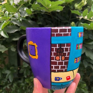 Peut inclure: Une tasse en c&eacute;ramique violette et bleue peinte avec une sc&egrave;ne de la s&eacute;rie t&eacute;l&eacute;vis&eacute;e "Friends". La tasse pr&eacute;sente un mur de briques, une fen&ecirc;tre, une porte et une biblioth&egrave;que. La tasse est tenue dans la main d'une personne.