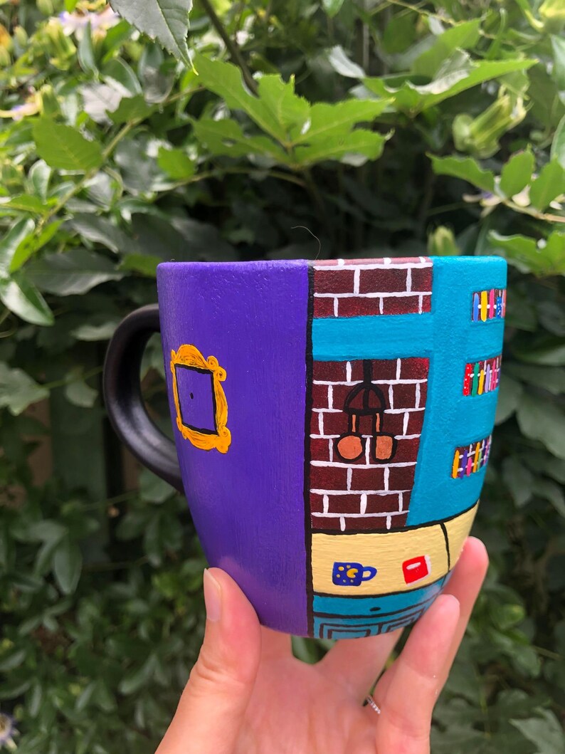 Peut inclure: Une tasse en c&eacute;ramique violette et bleue peinte avec une sc&egrave;ne de la s&eacute;rie t&eacute;l&eacute;vis&eacute;e "Friends". La tasse pr&eacute;sente un mur de briques, une fen&ecirc;tre, une porte et une biblioth&egrave;que. La tasse est tenue dans la main d'une personne.