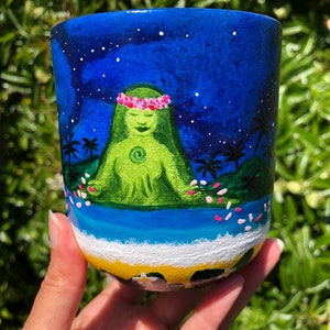 Pode incluir: Uma caneca de cer&acirc;mica pintada &agrave; m&atilde;o com um fundo de c&eacute;u noturno azul, estrelas brancas e uma figura verde com uma coroa de flores rosa. A figura est&aacute; sentada em uma ilha verde com palmeiras e flores rosas caindo do c&eacute;u. A parte inferior da caneca mostra um oceano azul com ondas brancas e uma praia amarela.