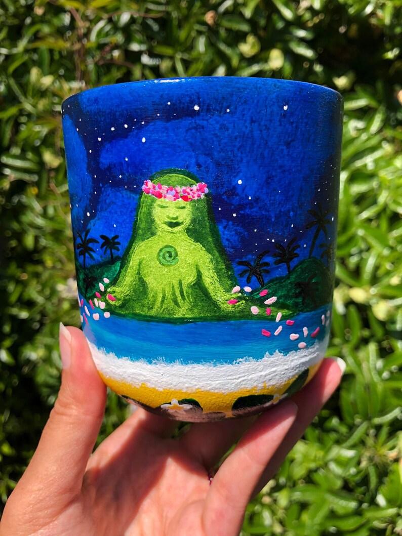 Pode incluir: Uma caneca de cer&acirc;mica pintada &agrave; m&atilde;o com um fundo de c&eacute;u noturno azul, estrelas brancas e uma figura verde com uma coroa de flores rosa. A figura est&aacute; sentada em uma ilha verde com palmeiras e flores rosas caindo do c&eacute;u. A parte inferior da caneca mostra um oceano azul com ondas brancas e uma praia amarela.