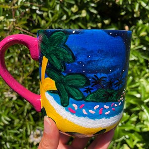 Pode incluir: Uma caneca de cer&acirc;mica pintada &agrave; m&atilde;o com uma cena de ilha tropical. A caneca apresenta um c&eacute;u azul com estrelas brancas, palmeiras, uma praia amarela e flores cor-de-rosa. A caneca tem uma al&ccedil;a rosa.
