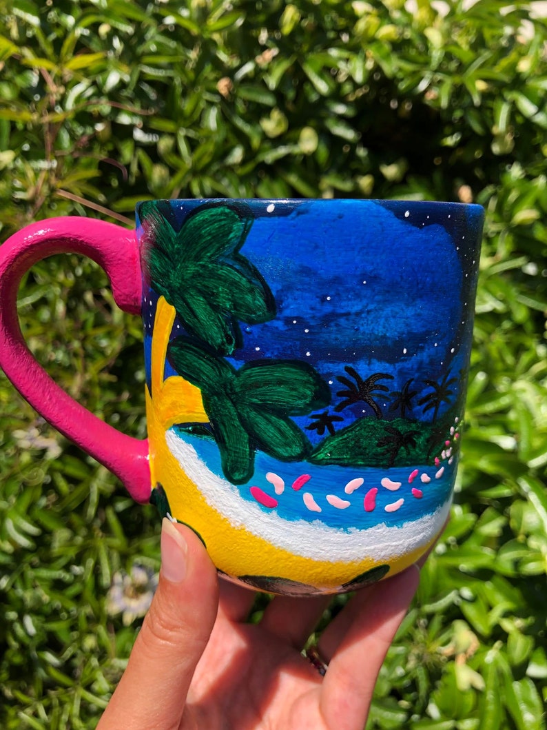Pode incluir: Uma caneca de cer&acirc;mica pintada &agrave; m&atilde;o com uma cena de ilha tropical. A caneca apresenta um c&eacute;u azul com estrelas brancas, palmeiras, uma praia amarela e flores cor-de-rosa. A caneca tem uma al&ccedil;a rosa.