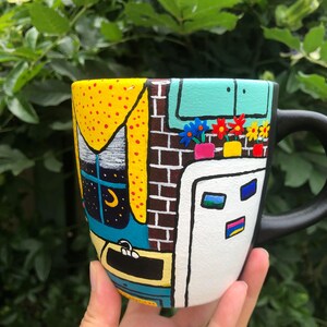 Peut inclure: Une tasse en c&eacute;ramique noire peinte avec une sc&egrave;ne color&eacute;e d'une maison avec une fen&ecirc;tre, un mur de briques, une porte et des fleurs. La tasse a une poign&eacute;e noire.