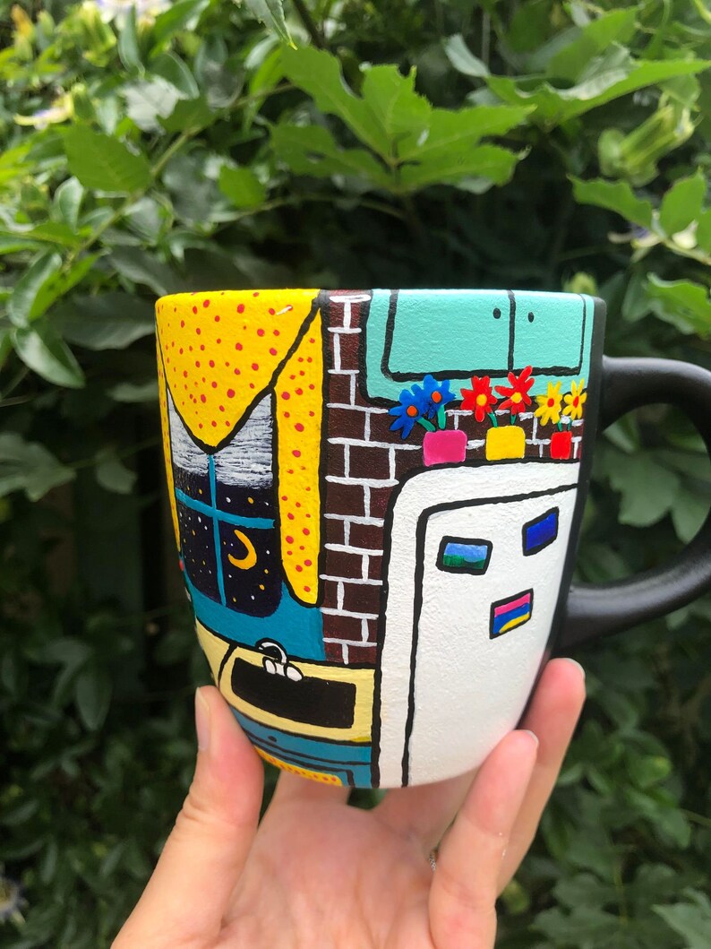 Peut inclure: Une tasse en c&eacute;ramique noire peinte avec une sc&egrave;ne color&eacute;e d'une maison avec une fen&ecirc;tre, un mur de briques, une porte et des fleurs. La tasse a une poign&eacute;e noire.