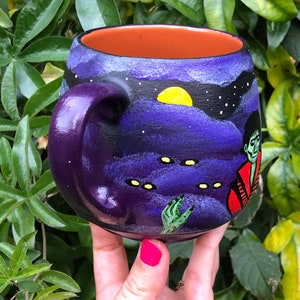 K&ouml;nnte beinhalten: Eine lila Keramiktasse mit einer handgemalten, gruseligen Szene. Das Design zeigt einen Nachthimmel mit gelbem Mond, Sternen und einem Zombie. Der Innenraum ist orange. Die Tasse wird vor einem Hintergrund aus gr&uuml;nen Bl&auml;ttern gehalten.