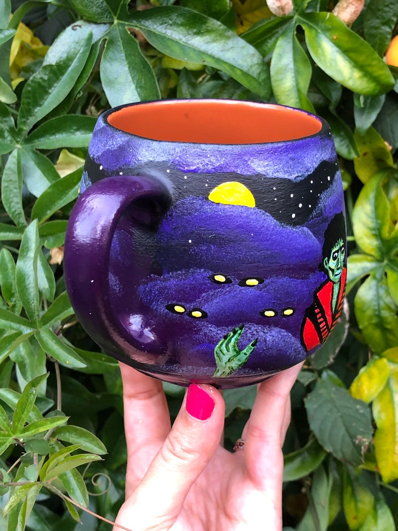 K&ouml;nnte beinhalten: Eine lila Keramiktasse mit einer handgemalten, gruseligen Szene. Das Design zeigt einen Nachthimmel mit gelbem Mond, Sternen und einem Zombie. Der Innenraum ist orange. Die Tasse wird vor einem Hintergrund aus gr&uuml;nen Bl&auml;ttern gehalten.