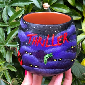 K&ouml;nnte beinhalten: Handbemalte Keramiktasse mit einem lila Nachthimmel-Design und dem Wort "THRILLER" in Rot. Mit einem Zombie und einer gr&uuml;nen Handgrafik. Die Tasse hat einen orangefarbenen Innenraum.