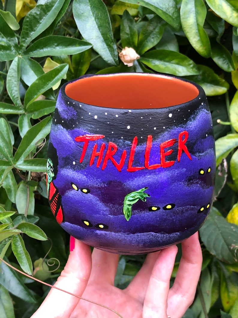 K&ouml;nnte beinhalten: Handbemalte Keramiktasse mit einem lila Nachthimmel-Design und dem Wort "THRILLER" in Rot. Mit einem Zombie und einer gr&uuml;nen Handgrafik. Die Tasse hat einen orangefarbenen Innenraum.