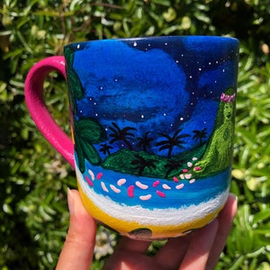 Pode incluir: Uma caneca de cer&acirc;mica rosa e branca com um design pintado &agrave; m&atilde;o de uma cena de ilha tropical. A caneca apresenta um c&eacute;u azul com estrelas brancas, palmeiras verdes, uma ilha verde com uma coroa de flores rosa e um oceano azul com ondas brancas e flores rosas.