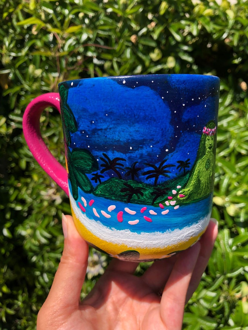 Pode incluir: Uma caneca de cer&acirc;mica rosa e branca com um design pintado &agrave; m&atilde;o de uma cena de ilha tropical. A caneca apresenta um c&eacute;u azul com estrelas brancas, palmeiras verdes, uma ilha verde com uma coroa de flores rosa e um oceano azul com ondas brancas e flores rosas.