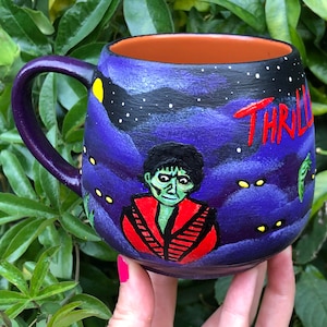 K&ouml;nnte beinhalten: Lila Keramikbecher mit einem bemalten Design mit einer zombieartigen Figur in einer roten Jacke, mit dem Wort "THRILL" in Rot. Der Becher hat einen lila Griff und eine dunkelorangefarbene Innenseite, vor einem sternenklaren Nachthimmel.