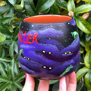 K&ouml;nnte beinhalten: Handbemalte Keramiktasse mit einem dunkelvioletten und schwarzen Nachthimmel-Design. Das Wort "KILLER" ist rot geschrieben. Die Tasse hat gelbe Augen und gr&uuml;ne, au&szlig;erirdische Figuren. Der Innenraum ist orange.