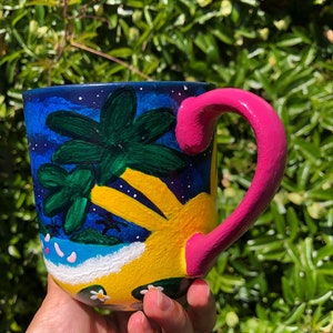 Pode incluir: Uma caneca de cer&acirc;mica pintada &agrave; m&atilde;o com uma cena tropical de cor amarela brilhante, azul e verde. A caneca tem uma al&ccedil;a rosa e flores brancas pintadas no lado.