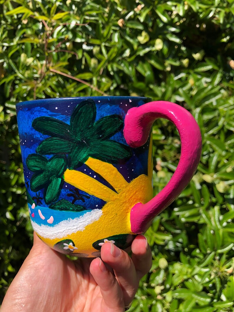 Pode incluir: Uma caneca de cer&acirc;mica pintada &agrave; m&atilde;o com uma cena tropical de cor amarela brilhante, azul e verde. A caneca tem uma al&ccedil;a rosa e flores brancas pintadas no lado.