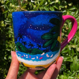 Pode incluir: Uma caneca de cer&acirc;mica azul com uma cena pintada &agrave; m&atilde;o de uma ilha tropical &agrave; noite. A caneca tem uma al&ccedil;a rosa e estrelas brancas pintadas no fundo azul. A cena da ilha inclui palmeiras, uma praia e um sol amarelo.