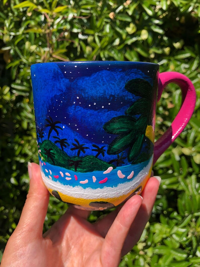 Pode incluir: Uma caneca de cer&acirc;mica azul com uma cena pintada &agrave; m&atilde;o de uma ilha tropical &agrave; noite. A caneca tem uma al&ccedil;a rosa e estrelas brancas pintadas no fundo azul. A cena da ilha inclui palmeiras, uma praia e um sol amarelo.