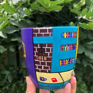 Peut inclure: Un pot en c&eacute;ramique peint &agrave; la main en forme de l'immeuble embl&eacute;matique de la s&eacute;rie t&eacute;l&eacute;vis&eacute;e "Friends". Le pot est peint dans des tons de violet, bleu, jaune et marron, avec des d&eacute;tails comme un mur en briques, une fen&ecirc;tre et une biblioth&egrave;que.