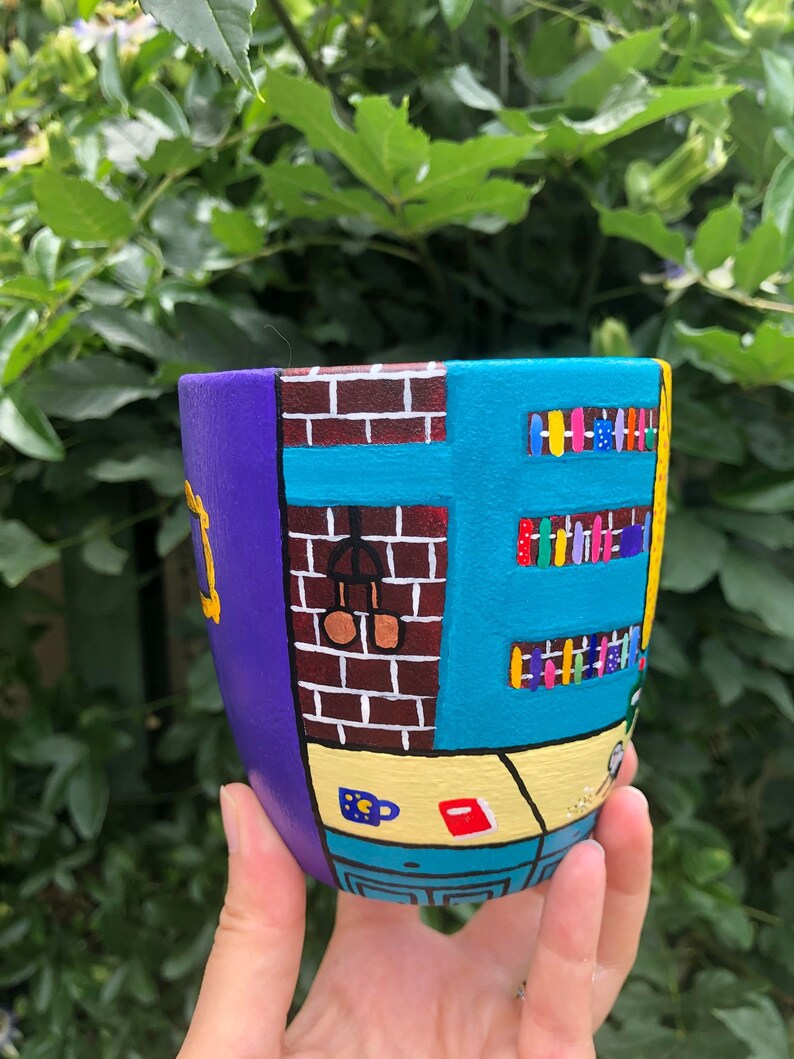 Peut inclure: Un pot en c&eacute;ramique peint &agrave; la main en forme de l'immeuble embl&eacute;matique de la s&eacute;rie t&eacute;l&eacute;vis&eacute;e "Friends". Le pot est peint dans des tons de violet, bleu, jaune et marron, avec des d&eacute;tails comme un mur en briques, une fen&ecirc;tre et une biblioth&egrave;que.