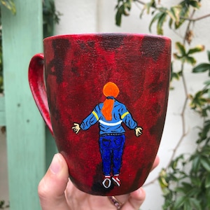 May include: A red ceramic mug with a hand-painted design of a person with red hair wearing a blue jacket and blue jeans. The person is facing away from the viewer.