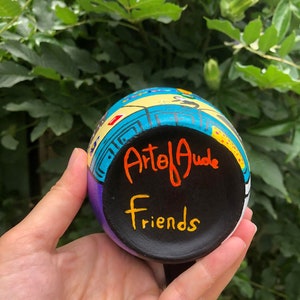 Peut inclure: Une tasse en c&eacute;ramique peinte &agrave; la main avec un motif color&eacute;. La tasse a une base noire avec les mots "Arte of Jude" et "Friends" &eacute;crits en peinture orange et jaune.