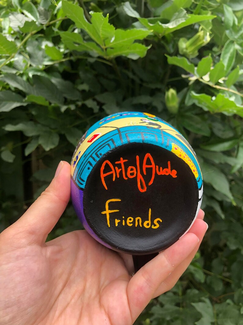 Peut inclure: Une tasse en c&eacute;ramique peinte &agrave; la main avec un motif color&eacute;. La tasse a une base noire avec les mots "Arte of Jude" et "Friends" &eacute;crits en peinture orange et jaune.