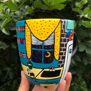 Peut inclure: Une tasse en c&eacute;ramique peinte avec une sc&egrave;ne fantaisiste d'un int&eacute;rieur de cuisine. La tasse pr&eacute;sente une fen&ecirc;tre jaune avec une lune croissante et des &eacute;toiles, un comptoir bleu et un &eacute;vier noir. La tasse est peinte dans un style cartoon avec des couleurs vives.