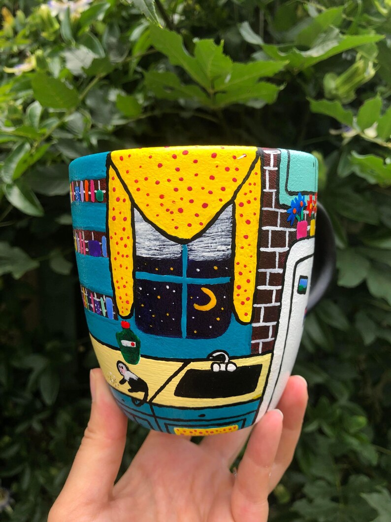 Peut inclure: Une tasse en c&eacute;ramique peinte avec une sc&egrave;ne fantaisiste d'un int&eacute;rieur de cuisine. La tasse pr&eacute;sente une fen&ecirc;tre jaune avec une lune croissante et des &eacute;toiles, un comptoir bleu et un &eacute;vier noir. La tasse est peinte dans un style cartoon avec des couleurs vives.