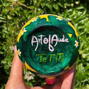 Pode incluir: Uma caneca de cer&acirc;mica amarela e verde com flores brancas e as palavras "Atol Auda Te Fiti" pintadas no lado.