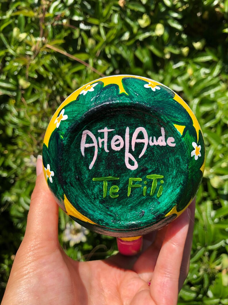 Pode incluir: Uma caneca de cer&acirc;mica amarela e verde com flores brancas e as palavras "Atol Auda Te Fiti" pintadas no lado.