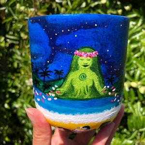 Pode incluir: Caneca de cer&acirc;mica pintada &agrave; m&atilde;o com um fundo de c&eacute;u noturno azul, estrelas brancas e uma figura verde com uma coroa de flores rosa sentada em posi&ccedil;&atilde;o de l&oacute;tus. A figura &eacute; cercada por flores rosas e palmeiras. O fundo da caneca &eacute; pintado com uma cena de praia amarela.