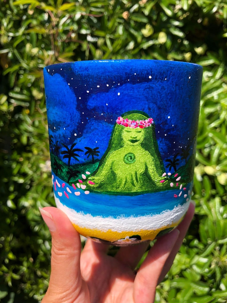 Pode incluir: Caneca de cer&acirc;mica pintada &agrave; m&atilde;o com um fundo de c&eacute;u noturno azul, estrelas brancas e uma figura verde com uma coroa de flores rosa sentada em posi&ccedil;&atilde;o de l&oacute;tus. A figura &eacute; cercada por flores rosas e palmeiras. O fundo da caneca &eacute; pintado com uma cena de praia amarela.