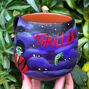 K&ouml;nnte beinhalten: Handbemalte Keramiktasse mit einem lila und schwarzen Nachthimmel-Design. Das Wort "THRILLER" ist rot geschrieben. Die Tasse zeigt einen Zombie und eine gr&uuml;ne Hand, die ein gruseliges Thema hervorrufen.