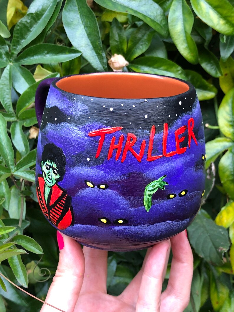 K&ouml;nnte beinhalten: Handbemalte Keramiktasse mit einem lila und schwarzen Nachthimmel-Design. Das Wort "THRILLER" ist rot geschrieben. Die Tasse zeigt einen Zombie und eine gr&uuml;ne Hand, die ein gruseliges Thema hervorrufen.