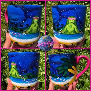Pode incluir: Quatro vistas diferentes de uma caneca de cer&acirc;mica branca com um design pintado &agrave; m&atilde;o. A caneca apresenta um c&eacute;u noturno azul com estrelas, uma ilha verde com uma mulher com uma coroa de flores e flores brancas caindo do c&eacute;u. A caneca tem uma al&ccedil;a rosa.