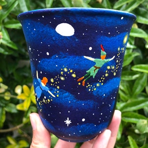 Peut inclure: Une tasse en céramique bleue avec une scène peinte à la main de Peter Pan et de la fée Clochette volant dans un ciel nocturne étoilé. La tasse présente une lune blanche, des étoiles jaunes et des nuages bleus.