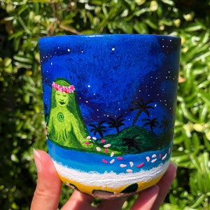 Pode incluir: Uma caneca de cer&acirc;mica pintada &agrave; m&atilde;o com um fundo de c&eacute;u noturno azul, estrelas brancas, uma ilha verde com palmeiras e uma figura verde com uma coroa de flores rosa. A base da caneca &eacute; pintada com uma praia amarela e um oceano azul.