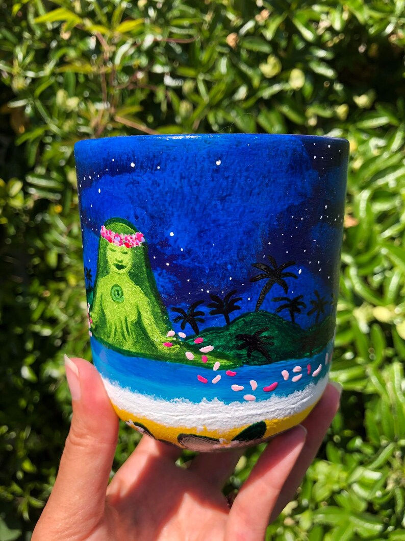 Pode incluir: Uma caneca de cer&acirc;mica pintada &agrave; m&atilde;o com um fundo de c&eacute;u noturno azul, estrelas brancas, uma ilha verde com palmeiras e uma figura verde com uma coroa de flores rosa. A base da caneca &eacute; pintada com uma praia amarela e um oceano azul.