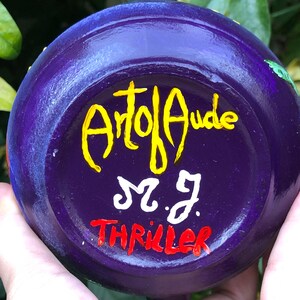 K&ouml;nnte beinhalten: Eine handbemalte, violette Keramikschale mit den Texten "Art of Aude", "M.J." und "Thriller" in Gelb, Wei&szlig; und Rot. Die Schale wird in der Hand einer Person gehalten, mit gr&uuml;nem Laub im Hintergrund.