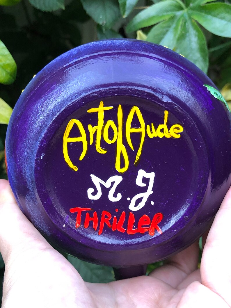K&ouml;nnte beinhalten: Eine handbemalte, violette Keramikschale mit den Texten "Art of Aude", "M.J." und "Thriller" in Gelb, Wei&szlig; und Rot. Die Schale wird in der Hand einer Person gehalten, mit gr&uuml;nem Laub im Hintergrund.