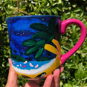 Pode incluir: Uma caneca de cer&acirc;mica pintada &agrave; m&atilde;o com uma cena de praia tropical. A caneca apresenta palmeiras, uma praia e ondas do oceano. A al&ccedil;a &eacute; rosa.