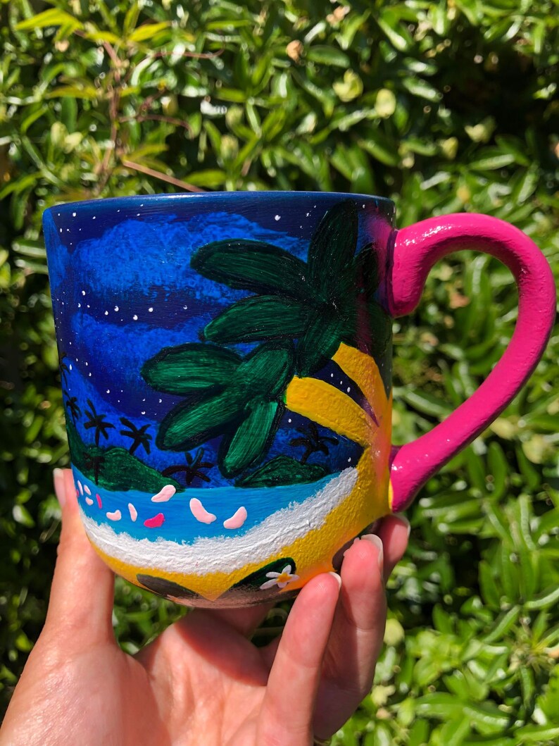 Pode incluir: Uma caneca de cer&acirc;mica pintada &agrave; m&atilde;o com uma cena de praia tropical. A caneca apresenta palmeiras, uma praia e ondas do oceano. A al&ccedil;a &eacute; rosa.